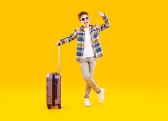 Excited tourist boy traveling with luggage on summer vacation. Happy caucasian teenager wearing sunglasses and trendy, stylish outfit standing against isolated yellow studio background