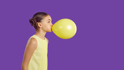 Cheerful funny little girl inflates yellow balloon isolated on vivid purple background. Profile...