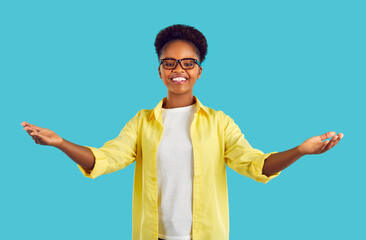 Happy cheerful afroamerican young woman with ponytail hair in white casual t-shirt and yellow shirt in glasses spreading arms wide ready to give a hug on turquoise background. Banner with copyspace.