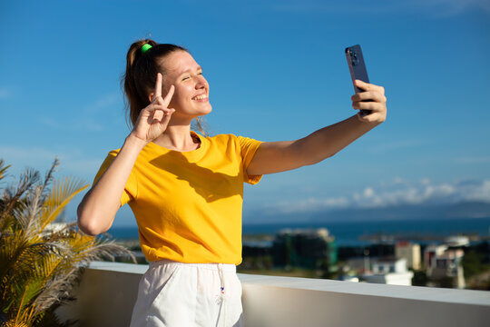 young beautiful happy woman blogger is taking selfie, picture of herself on frontal camera to social media of smartphone, cell mobile phone, smiling waving her hand, having video call, conversation