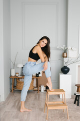 Young stylish brunette with long hair in a casual style, in jeans, a black knitted top, put her foot on a stepladder in the living room