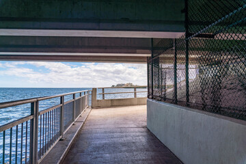 Concrete walkway at Rickenbacker Causeway underneath the bridge in Miami, Florida. Pathway with chainlink fence under a bridge over the water with views of cloudy sky at the background