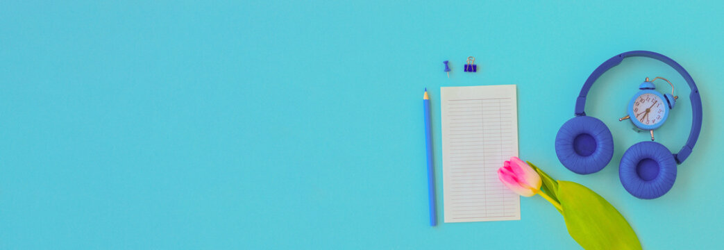 Banner flat lay home office desk. Workspace with pink tulip, headphones, alarm clock, pencil and a piece of paper for notes on blue background. Top view feminine background.