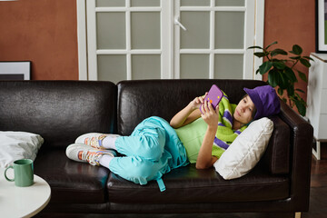 Teenage girl lying on sofa in living room and playing online games on her smartphone