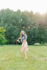 Naklejka na ściany i meble Young pretty woman in a summer dress walks through a meadow on green grass. A young girl holds shoes in her hands, showing off her beautiful leg. Summer outdoor recreation
