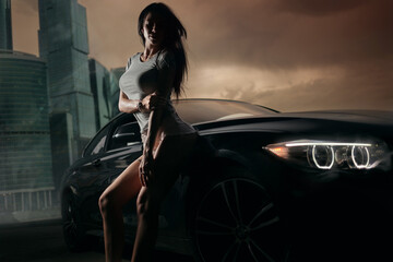Fototapeta na wymiar Attractive young sporty woman silhouette near a modern coupe cat in the evening in front of the city landscape with skyscrapers