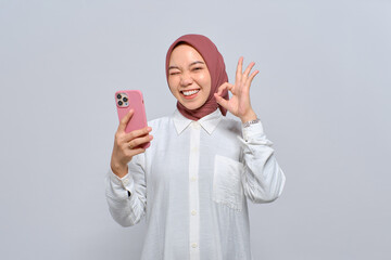 Smiling young Asian Muslim woman holding mobile phone and showing okay sign isolated over white...