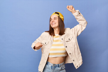 Image of attractive positive charming Caucasian teenager girl wearing baseball cap and jacket posing isolated over blue background, dancing at part, celebrating holiday.