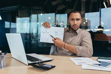 Fototapeta na wymiar Portrait disappointed businessman inside office, african american man showing document graph with bad financial performance to camera, financier in overcoat working inside office using laptop.