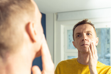 Obraz premium Mature man apply face tonic with cotton pad, then massage with hands. Male portrait in mirror. Skincare morning routine, careful beauty treatment, home lifestyle. Face cleansing, washing.