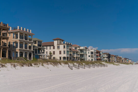 Three-storey beach houses with footbridges over sand dunes at Destin, Florida. Coastline residences with white sand and grass front views against the blue sky.