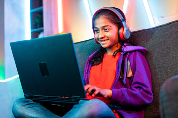 Happy teenager girl with headphones playing online video game on laptop while sitting on sofa at...