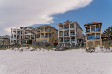 Facade of houses with footbridge access to the white sand beach in Destin, Florida. Multi-storey beach houses with sand dunes and grasses at the front against the giant clouds background.