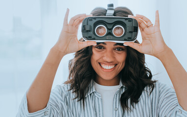 Young African American playful woman in casual shirt putting on vr headset looks at camera toothy smiles enjoying weekend home. Brazilian girl loves games got virtual reality googles on birthday.