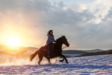 Romantic equestrian scene: A young woman ride her horse in gallop through a snowy winter landscape during sundown outdoors
