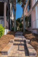 Narrow bricks path with steps in the middle of two houses near Four Prong Lake in Destin, Florida. Walkway with two palm trees and dried grasses at the side of the house buildings in a vertical shot.