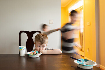 Sad Boy Sits Staring at Table as Brothers Run Around Behind Him