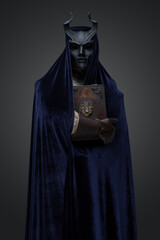 Shot of dark cultist dressed in horned mask and robe holding book.