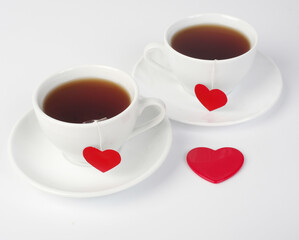Two white cups of tea on saucers with two red heart labels. White background.