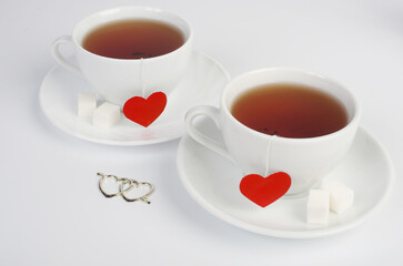 Two white cups of tea on saucers with two red heart labels. White background.
