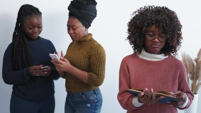 Outcast person. Social life. Friends jealousy. Happy women making selfie together while upset female with book staring them in light room interior.