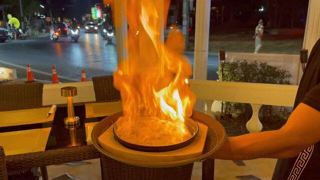 Anonymous waiter sets up fire Saganaki flaming cheese with torch at traditional Greek restaurant terrace at night
