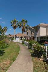 Obraz premium Concrete walkway on a grass outside the picket fence of houses in Navarre, Florida. Vertical shot view of pathway outside the residences with white picket fence along with the bushes and trees.