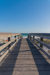 Wooden pathway overlooking ocean and blue sky on a sunny day in Destin Florida. The sunlit walkway leads to the sandy shore and sea with view of the horizon in the distance.
