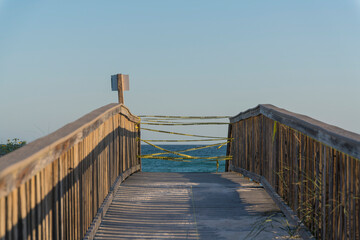 Naklejka premium Wooden sloped pathway with yellow caution tapes tied on the wooden railings in Destin, Florida. Boardwalk with views of the blue ocean and clear sky at the background.