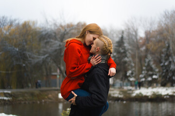 A guy with blond hair holds a red-haired girl in a red sweater in his arms near a winter lake, valentine's day