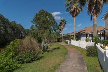 Obraz premium Concrete walkway on a grass with bushes outside the fenced residences in Navarre, Florida. There are plants and forest with tall trees on the left near the houses with white picket fence on the right.