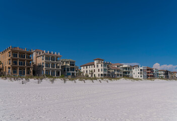 Fototapeta na wymiar Beach houses with roof decks and balconies facing the shore of the beach in Destin, Florida. There are fences of sand dunes with grasses with wooden footbridges at front of houses against the sky.