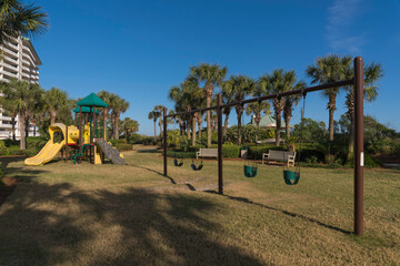 Playground in a park with benches, bushes, and palm trees near apartments. Views of slides against the building on the left and swings on the right against the trees and blue sky bacckground.