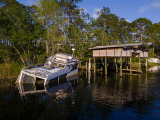 Half-sunken boat and wooden boat lift at the shore with tall trees and grasses in Navarre, Florida. There is a sunken boat on the left beside the boat lift on the right against the trees at the back.