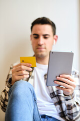 Young man shopping and paying online on digital tablet with credit card while relaxing at home listening music with earphones.