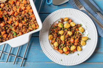 Ground beef stew. Cooked with peas and carrots, tomatoes, onions and herbs. Served with pan fried gnocchi on a plate on blue background. Flat lay