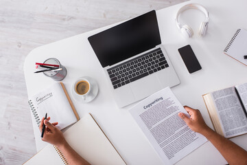 Top view of copywriter holding pen and papers near book and gadgets on table.