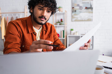 Indian copywriter holding paper near blurred laptop and cup of coffee at home.