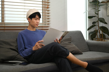 Peaceful young asian man relaxing on couch and reading book, spending leisure time at home