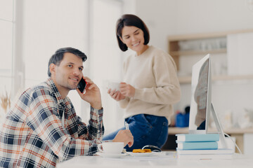 Photo of cheerful busy male freelancer uses mobile phone, discusses latest news, sits at desktop with papers and modern computer. Positive woman dressed in sweater and jeans, holds mug of drink