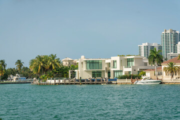 Fototapeta na wymiar Villas with private docks and palm trees outdoors in Miami, Florida bay. There is a modern white building with tall glass windows at the center and view of a high rise buildings at the back on right.