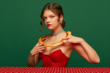 Lady. Young stylish beautiful girl with pizza crust on her shoulders sitting at table over green background. Vintage, retro style. Food pop art photography.