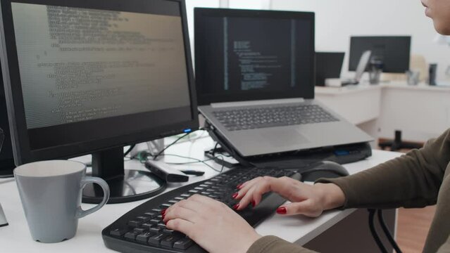Unrecognizable young woman sitting at desk with desktop computer and laptop on it writing code in programming language