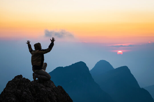 Silhouette Man kneeling down with hands open palm up praying to God on top mountain sunset background.