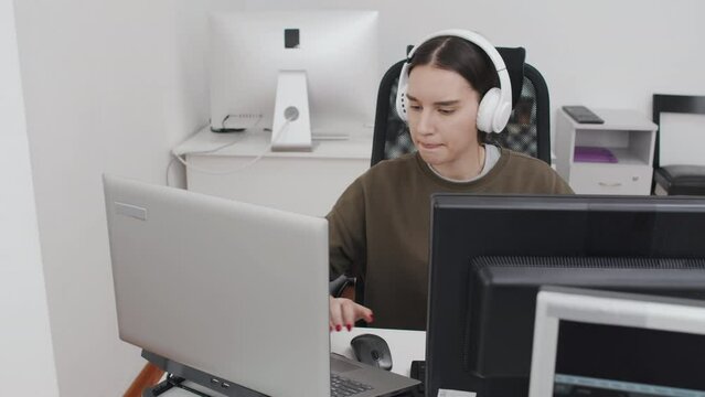 High angle of young Caucasian female IT engineer wearing wireless headphones decoding data using desktop pc and laptop