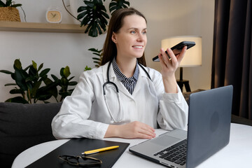 Smiling beautiful young woman doctor recording audio message on smartphone sitting at table with laptop, chatting with patient online, giving consultation or advice, treatment, using virtual voice app
