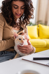 Smiling woman petting oriental cat near blurred notebook on table.