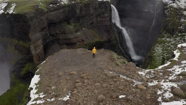 Aerial: One person near a cliff, wearing yellow jacket, walks towards a waterfall in Iceland