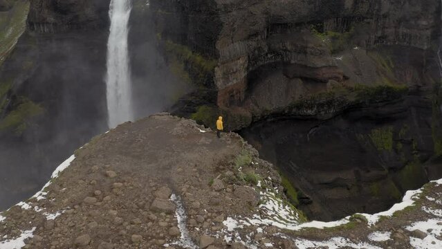 Aerial: Slow panning shot of a man standing near the edge of a cliff, watching Haifoss waterfall in Iceland