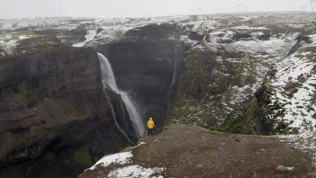 Aerial: Slow panning shot of a man standing near the edge of a cliff, watching Granni waterfall in Iceland